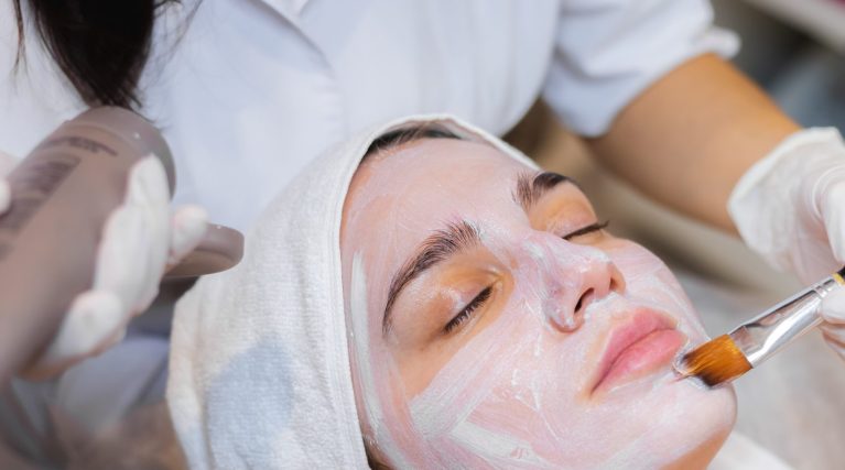 Beautician with a brush applies a white moisturizing mask to the face of a young girl client in a spa beauty salon